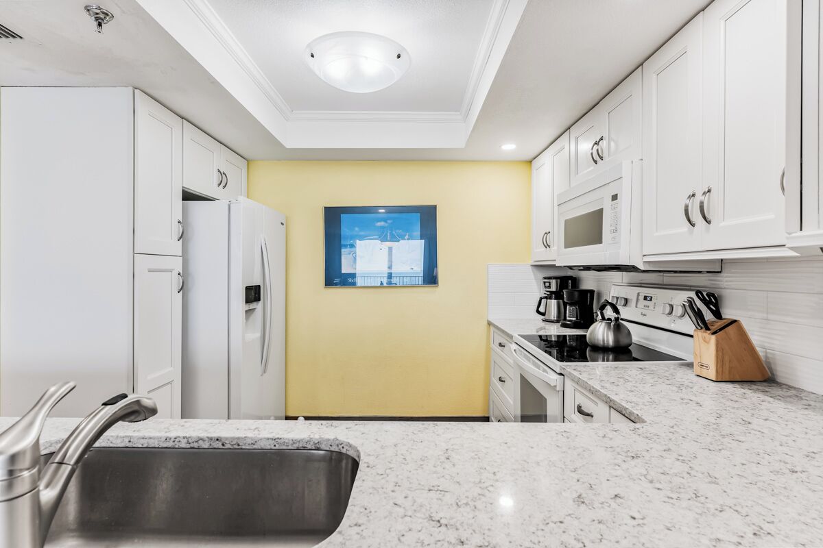 A modern kitchen with white cabinets, a yellow accent wall, a stainless steel sink, and a kettle on the stove.