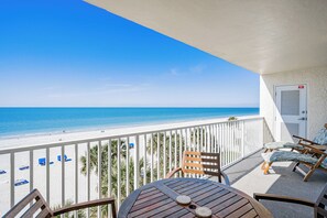 A balcony with a view of the beach, a round wooden table, two wooden chairs, and a door with a red sign.