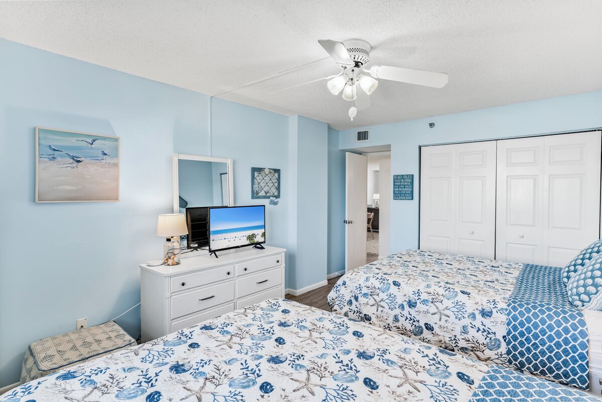 A bedroom with two beds, a TV on a dresser, a ceiling fan, and a door leading to another room.