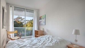 Un dormitorio con una cama grande, una silla de madera, una mesita con una lámpara, una ventana con cortinas y un cuadro en la pared.