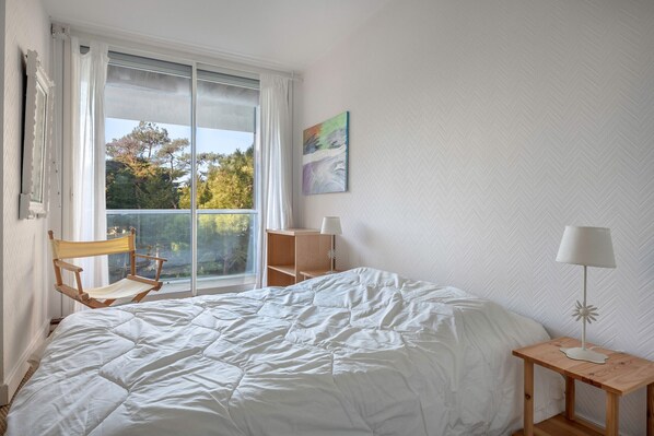 A bedroom with a large bed, a wooden chair, a small table with a lamp, a window with curtains, and a wall-mounted picture.
