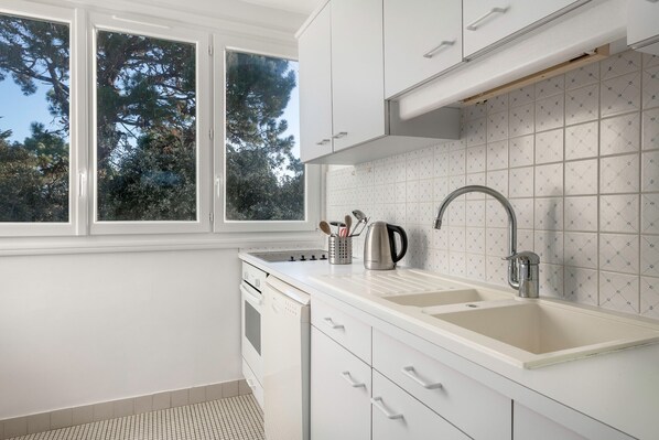 A modern kitchen with white cabinetry, a double sink, and a window overlooking trees.