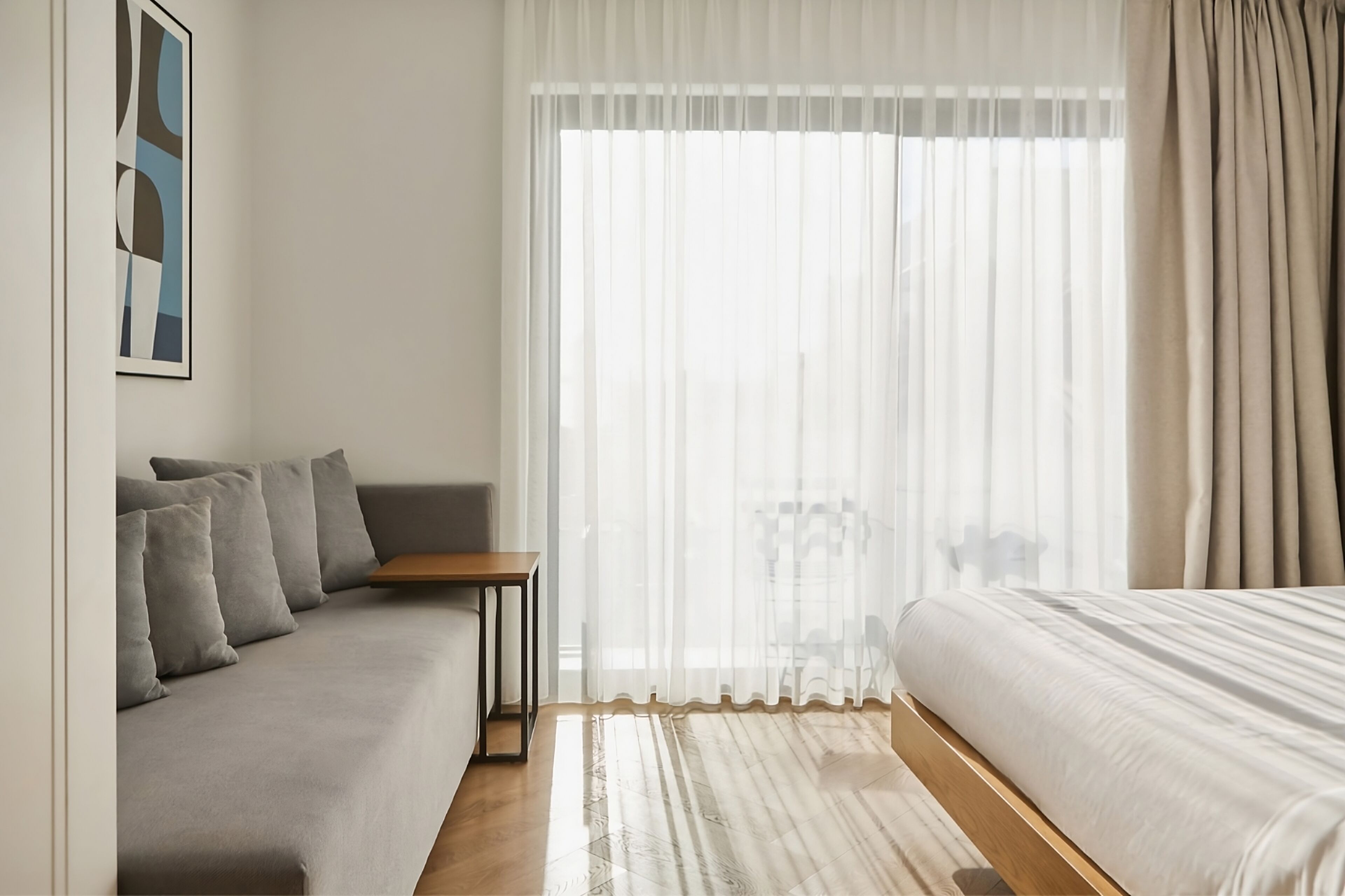Une chambre d’hôtel moderne avec un canapé gris, une table en bois, un lit avec une literie blanche et des rideaux transparents qui laissent passer la lumière naturelle.