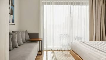 Une chambre d’hôtel moderne avec un canapé gris, une table en bois, un lit avec une literie blanche et des rideaux transparents qui laissent passer la lumière naturelle.