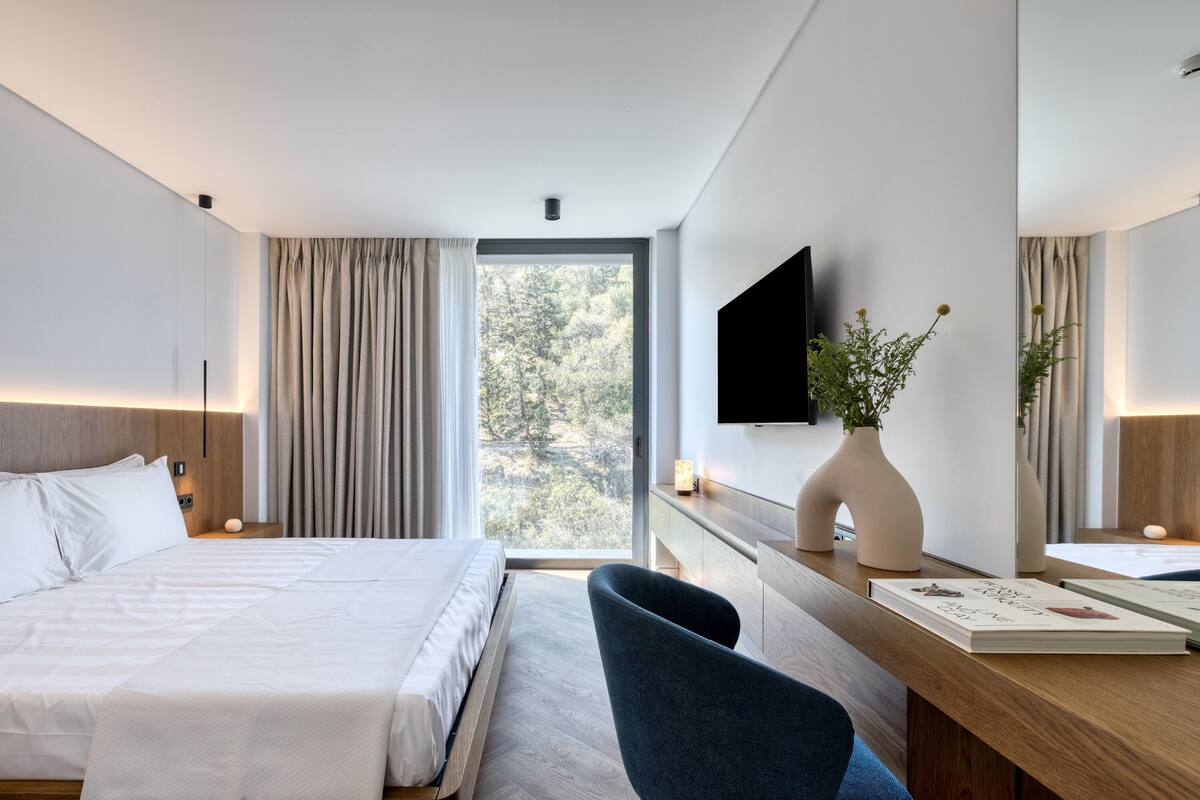 A modern hotel room with a large bed, a desk with a chair, a flat-screen TV, a vase with flowers, and a view of trees through the window.