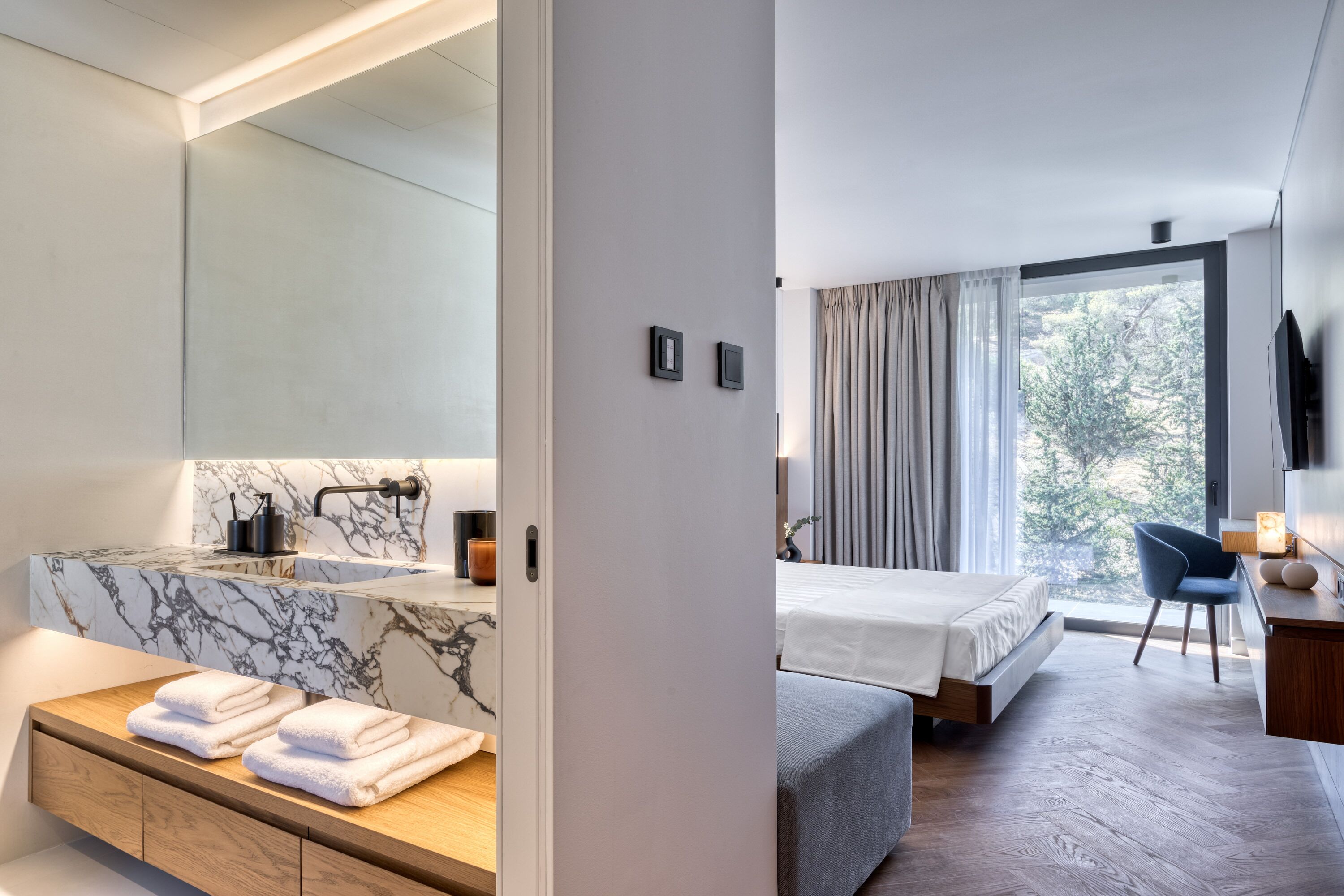 Une chambre d’hôtel moderne avec une salle de bain en marbre, un grand lit et une vue sur les arbres par la fenêtre.