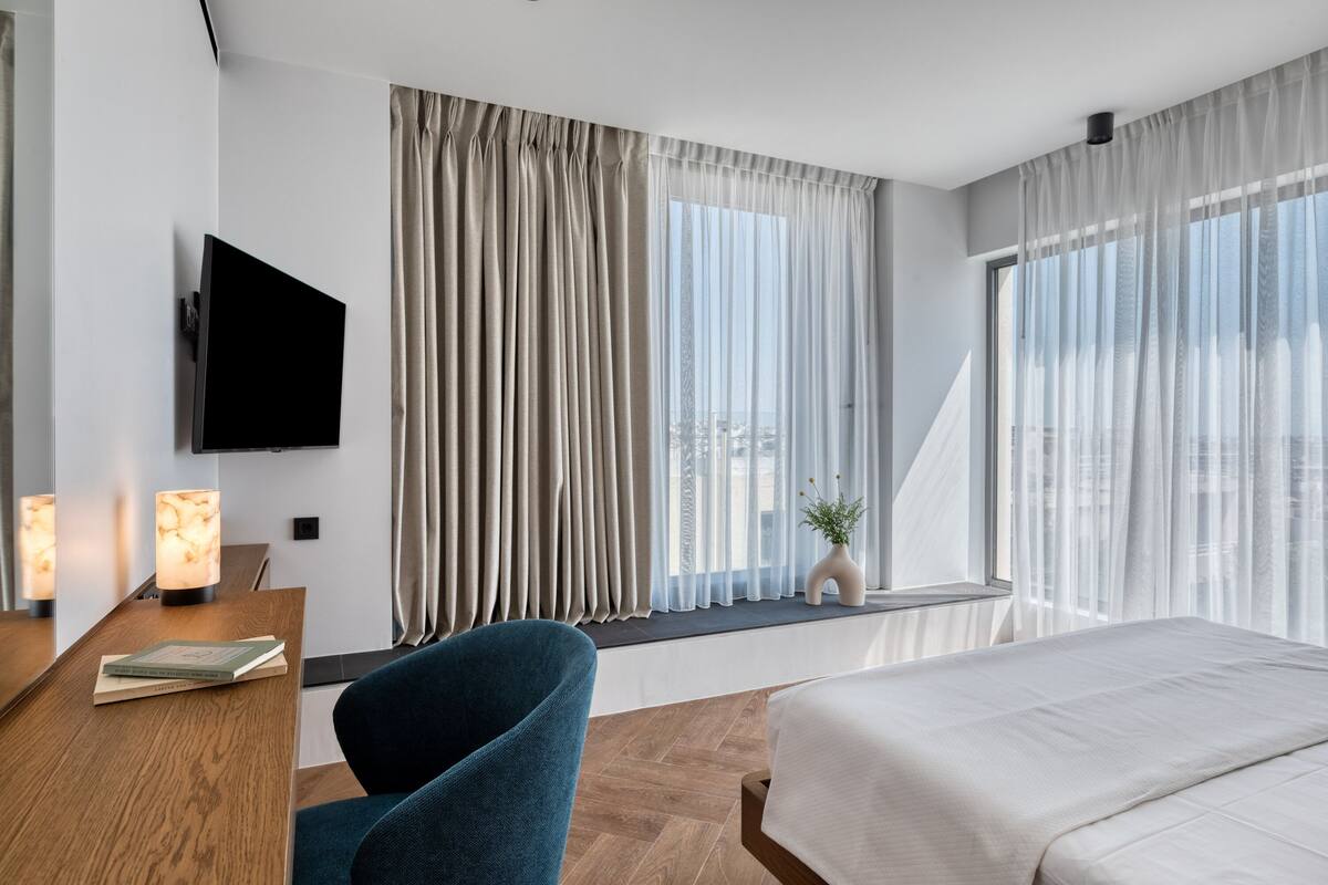 A modern hotel room with a large bed, a desk with a flat-screen TV, a chair, and a window with sheer curtains.
