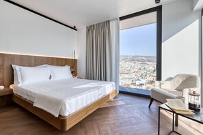 Une chambre d’hôtel moderne avec un grand lit, une tête de lit en bois, une vue sur le paysage urbain à travers une grande fenêtre, un coin salon avec un fauteuil et une table, et une petite bibliothèque.