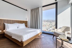 Moderni hotellihuone, jossa on suuri sänky, puinen pääty, näkymä kaupunkimaisemaan suuresta ikkunasta, oleskelualue tuolilla ja pöydällä sekä pieni kirjahylly.
