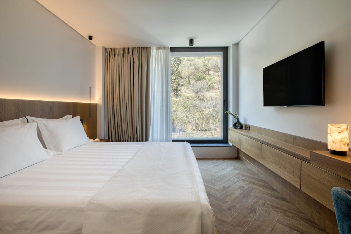 A modern hotel room with a large bed, a flat-screen TV mounted on the wall, a bedside table with a lamp, and a window with curtains.