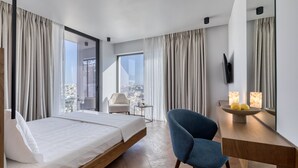 A modern hotel room with a bed, a desk with a bowl of fruit, a chair, and a view of the city. - Neoma  (Athens)