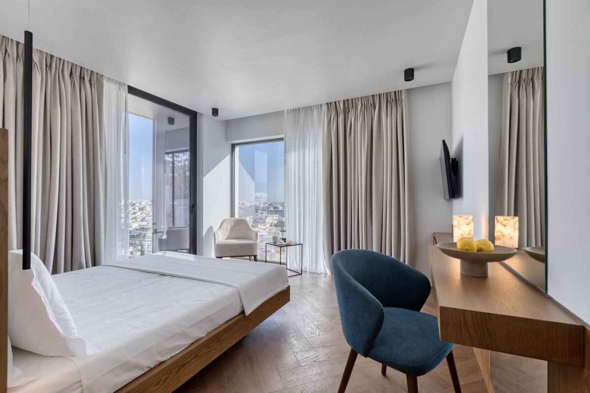 A modern hotel room with a bed, a desk with a bowl of fruit, a chair, and a view of the city.