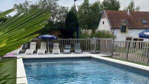 Una piscina rectangular con agua azul cristalina, rodeada por una valla de madera. Hay sillas de descanso y una sombrilla azul al borde de la piscina.