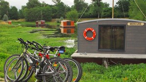 Una pequeña embarcación llamada 'Mercure' amarrada cerca de una zona con césped y bicicletas aparcadas.
