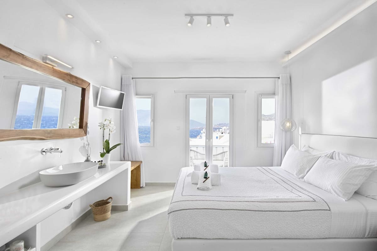A modern hotel room with a large bed, a vanity with a mirror, a sink, and a view of the sea through the windows.
