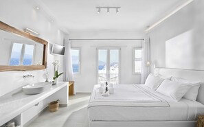Ein modernes Hotelzimmer mit einem großen Bett, einem Schminktisch mit Spiegel, einem Waschbecken und Meerblick durch die Fenster.