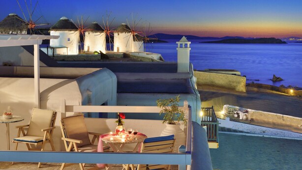 A seaside hotel with a terrace, lounge chairs, a table with drinks, and windmills in the background. - Marietta's Mykonos (Mykonos)