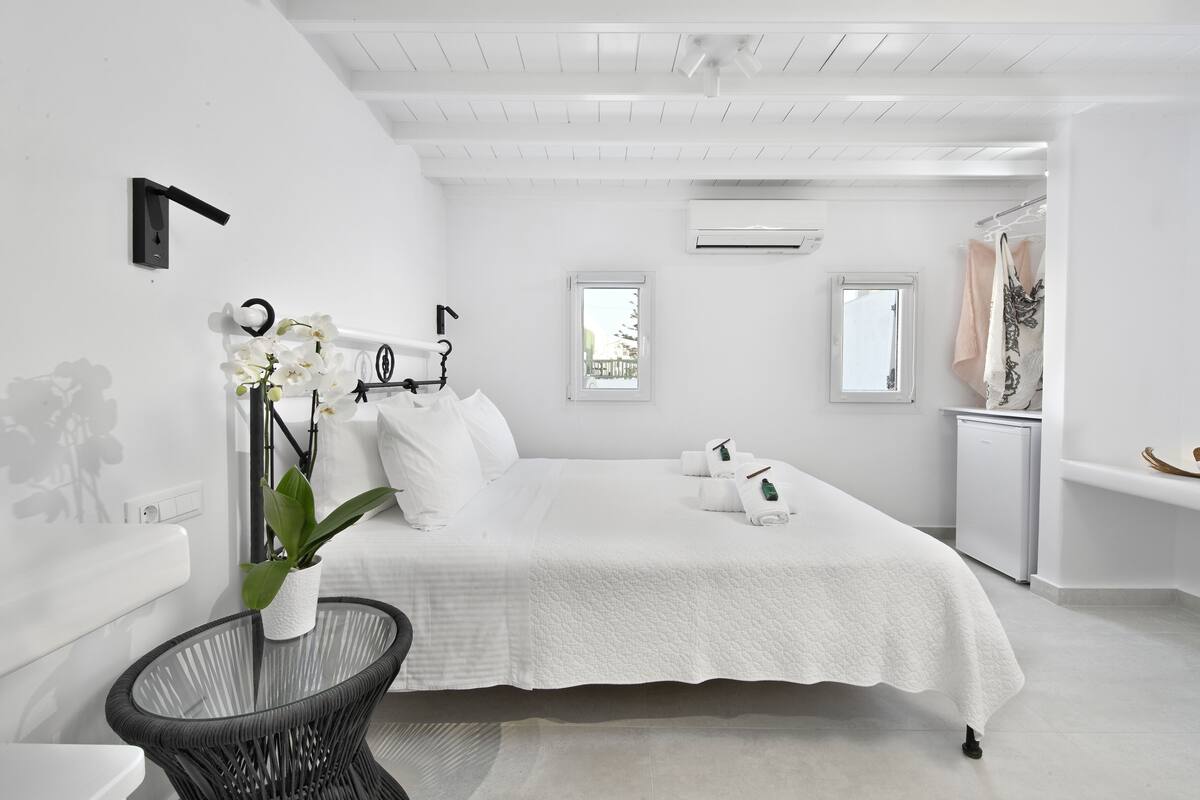 A white bedroom with a bed, two windows, a small table with a plant, and a white wardrobe.