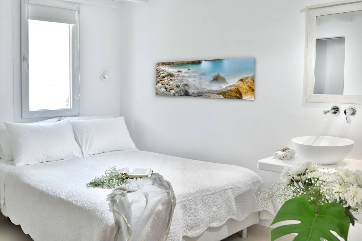 A bedroom with a bed, a bedside table with a bowl, a window, and a painting of a beach scene.
