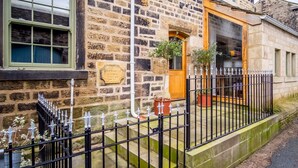 A stone building with a plaque, a wooden door, and a black metal fence. - School Cottage (Holmfirth)