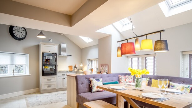 A modern kitchen with a built-in oven, a dining area with a wooden table and bench, and a large window with a view of the sky. - School Cottage (Holmfirth)