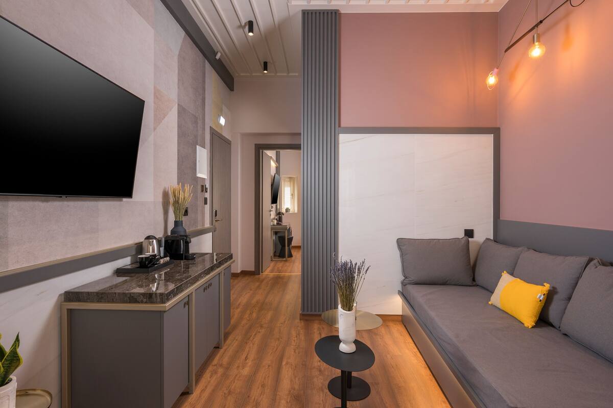 Una sala de estar moderna con un televisor de pantalla plana, un sofá con cojines, una mesita con un florero y una cocina al fondo.