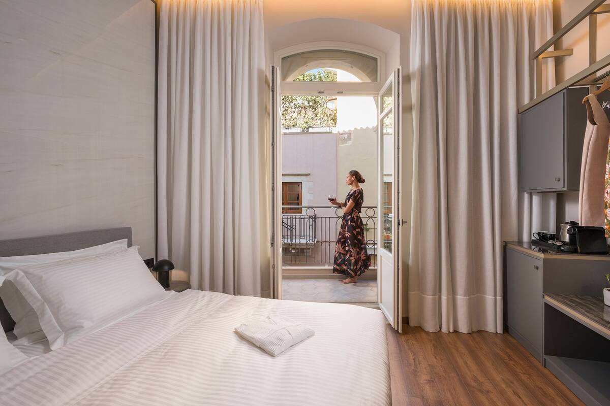 Habitación de hotel con cama, televisión, pequeña kitchenette y balcón con vistas al exterior.
