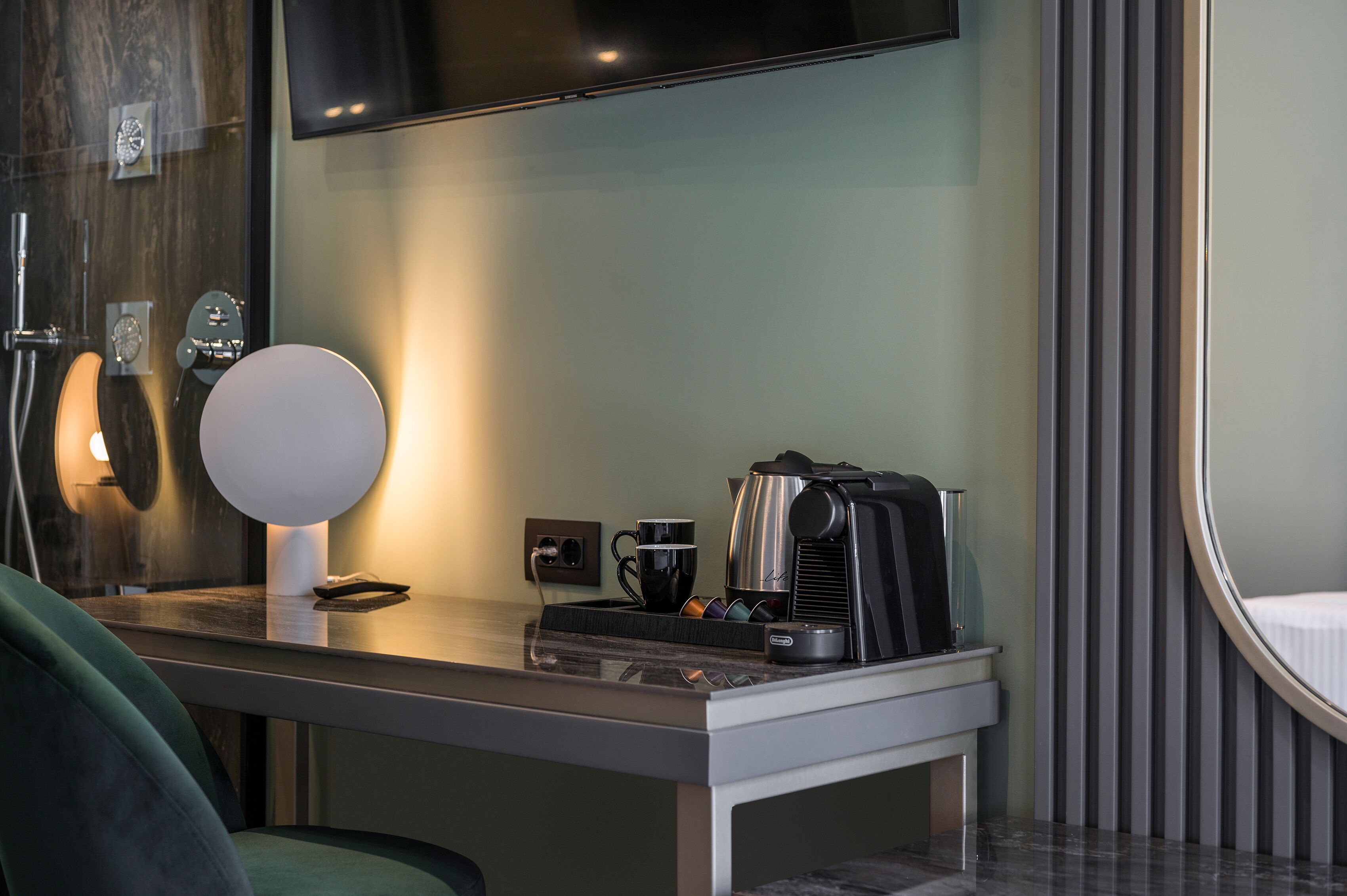 A modern hotel room with a sleek desk, a coffee maker, a cup, and a lamp.
