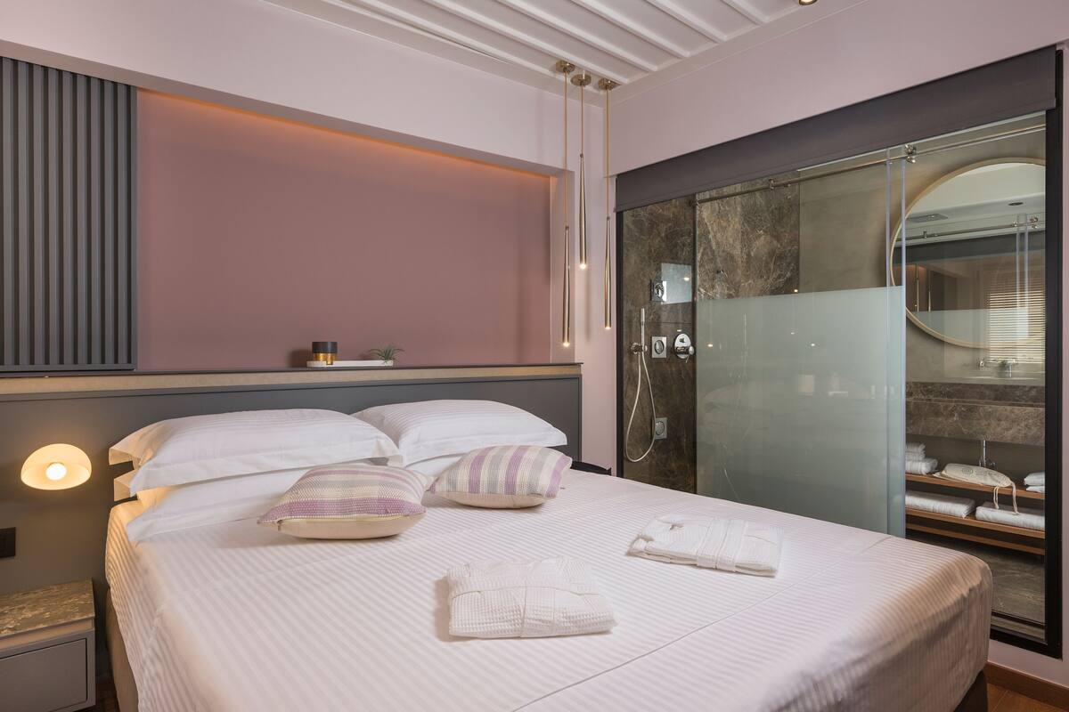 Habitación de hotel con cama, almohadas, mesita de noche, cabina de ducha de vidrio y vista al baño.