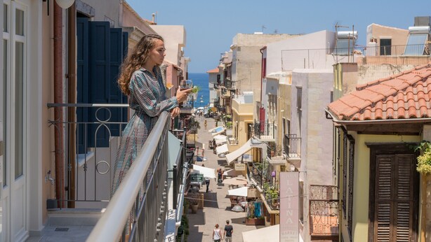 A balcony overlooking a narrow street with shops and buildings, a clear sky, and the sea in the distance. - Veneri Boutique Suites (Chania)