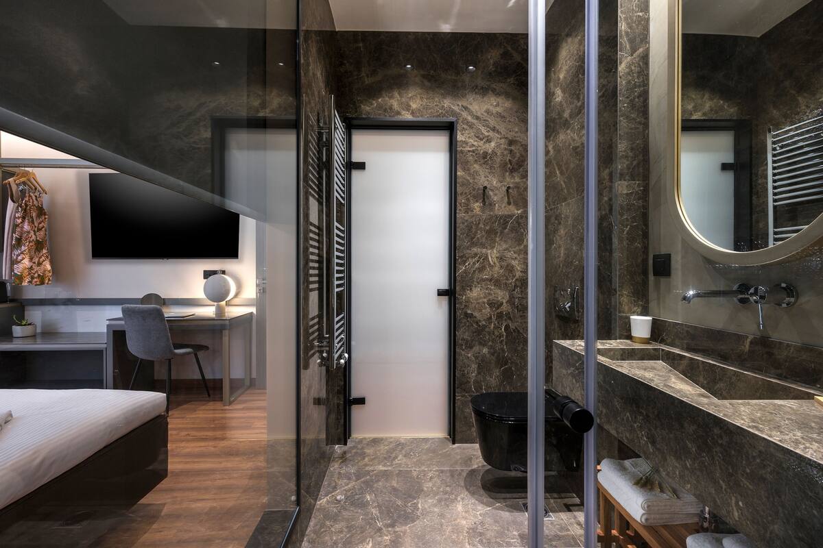 Un dormitorio moderno con cama, escritorio y silla, un baño con lavamanos y espejo, y zona de ducha.