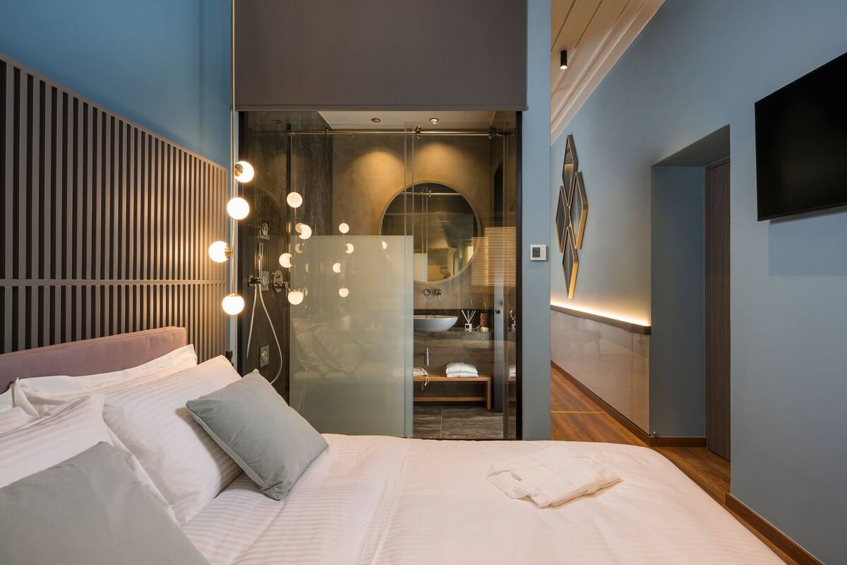 Una habitación de hotel moderna con cama, baño con paredes de vidrio y televisor montado.