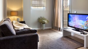 Ein Wohnzimmer mit einem dunkelgrauen Sofa, einem Flachbildfernseher auf einem weißen Schrank, einer Topfpflanze und einem Fenster mit Vorhängen.