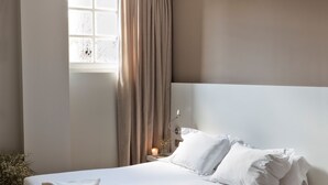 Un lit bien fait, avec du linge de lit blanc, une table de chevet avec une lampe et une chaise dans une pièce au plancher en bois et dotée d’une fenêtre avec des rideaux.