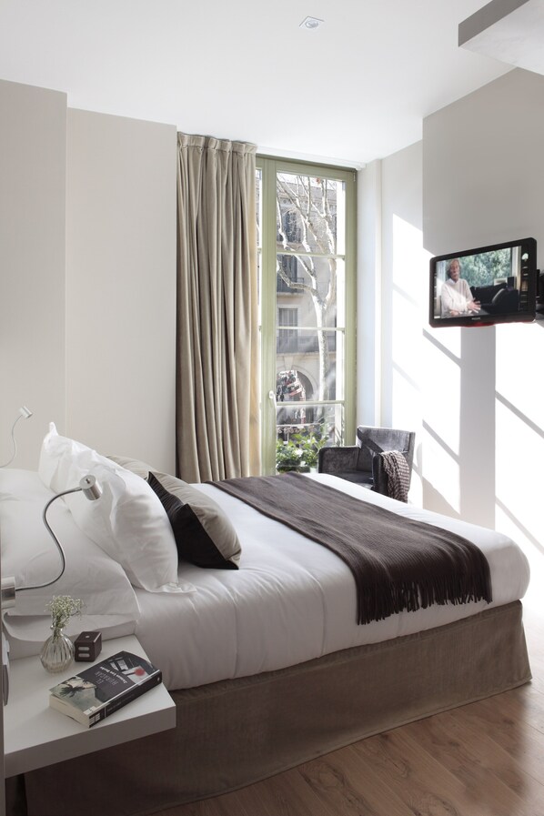 Een moderne hotelkamer met een groot bed, een nachtkastje met een boek, een aan de muur bevestigde televisie en een raam met gordijnen.