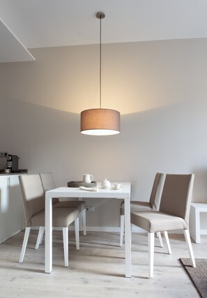 Een moderne eetruimte met een witte tafel en stoelen, een hanglamp en een koffieapparaat aan de muur.