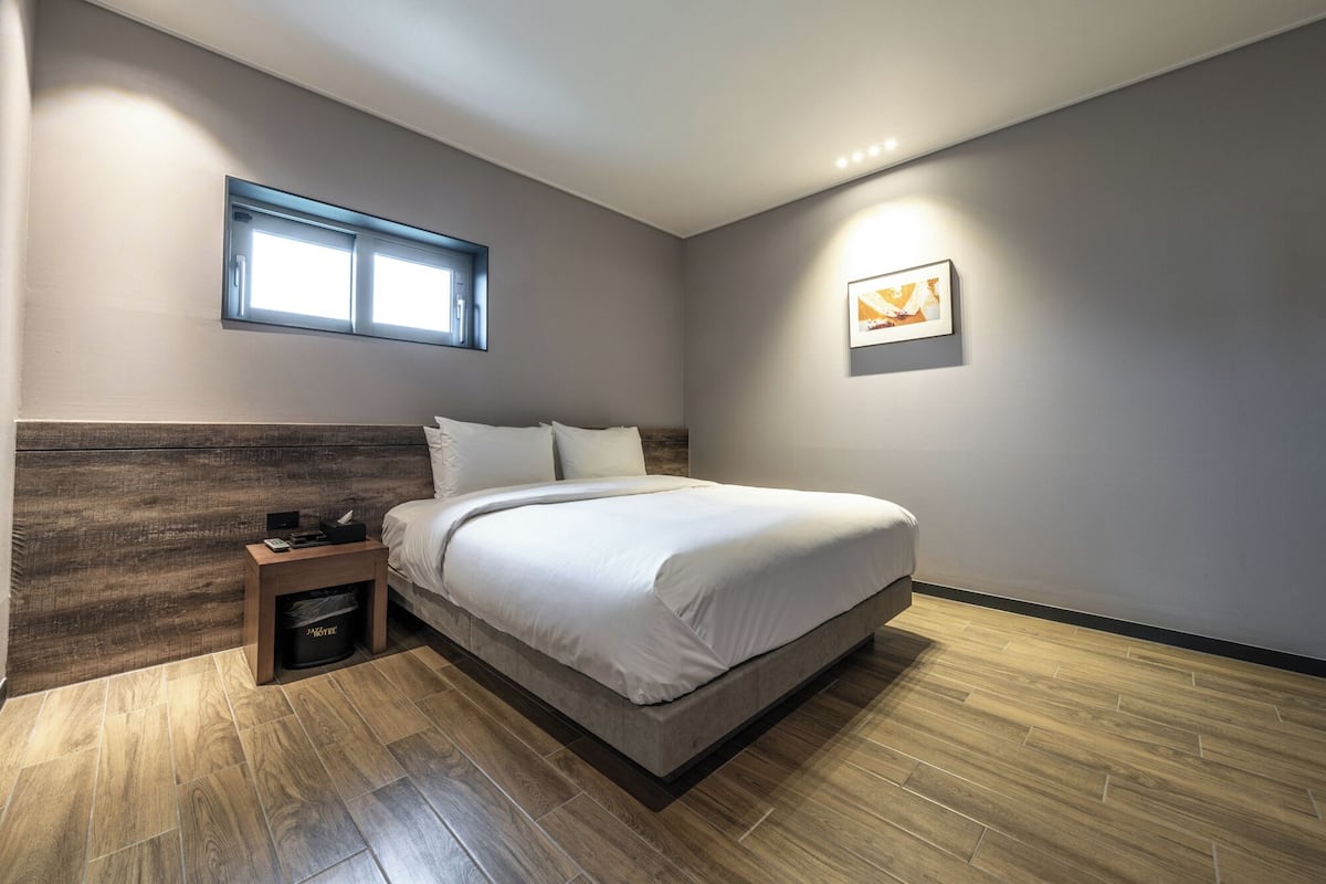 A modern bedroom with a bed, a nightstand, a trash bin, a window, and a wall-mounted picture.