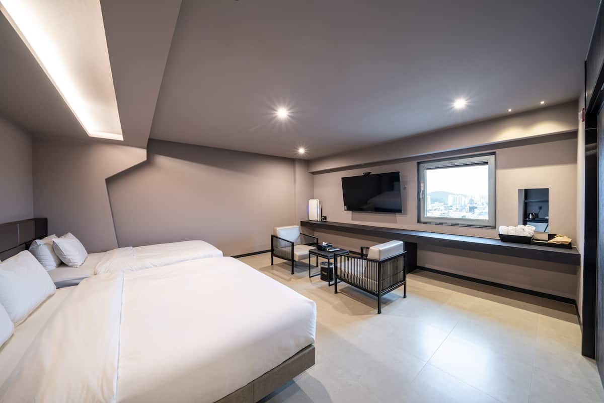 A modern hotel room with a large bed, a desk with a chair, a flat-screen TV mounted on the wall, and a window with a city view.
