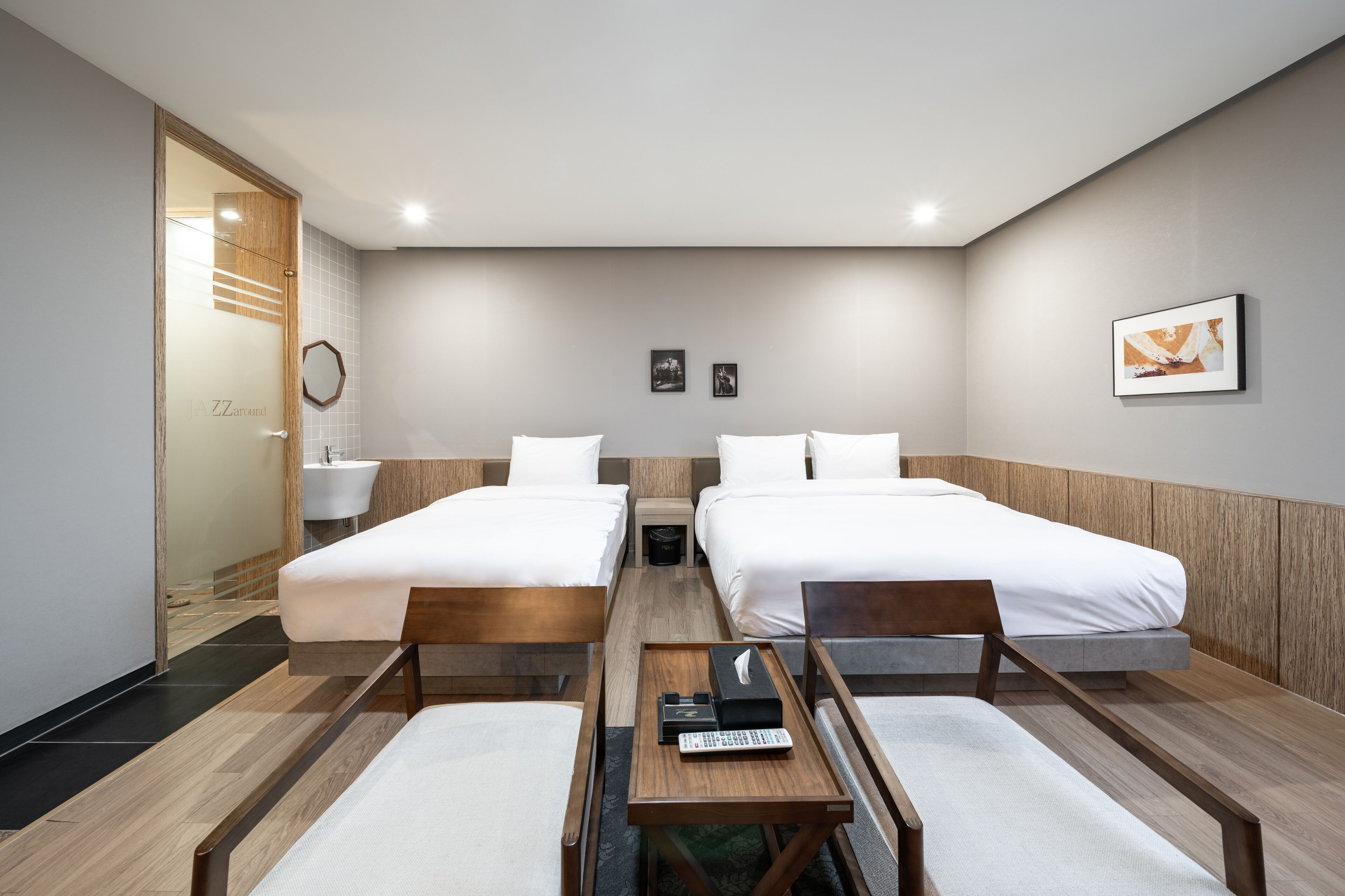 A hotel room with two beds, a wooden bench, a small table with a remote control, and a wall-mounted mirror.