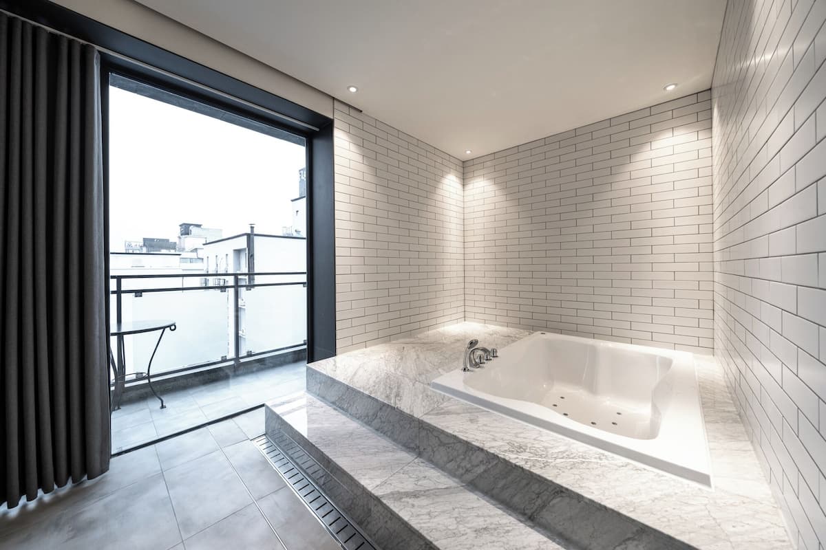 A modern bathroom with a large bathtub, white tiled walls, and a view through a large window.