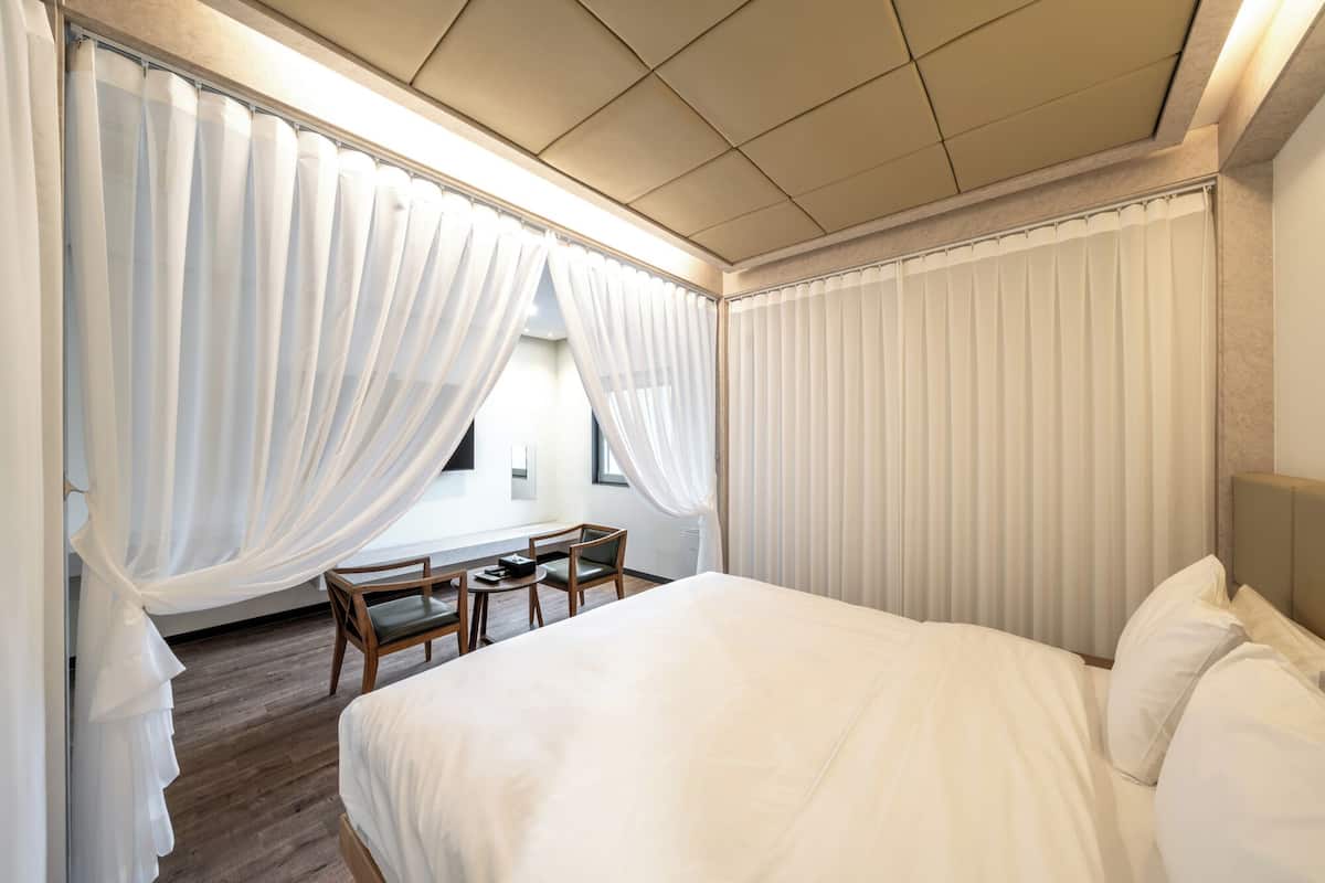 A hotel room with a bed, a small table, two chairs, and sheer curtains.