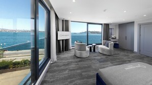 A modern hotel room with a large window offering a view of the sea and a cityscape. There are two sofas and a small table in the room. - Pera Bosphorus Hotel (Istanbul)