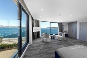 A modern hotel room with a large window offering a view of the sea and a cityscape. There are two sofas and a small table in the room.