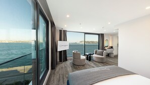 A modern hotel room with a large window offering a sea view, a comfortable bed, a seating area with a small table, and a vanity with a mirror. - Pera Bosphorus Hotel (Istanbul)