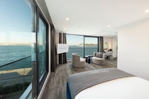 A modern hotel room with a large window offering a sea view, a comfortable bed, a seating area with a small table, and a vanity with a mirror.