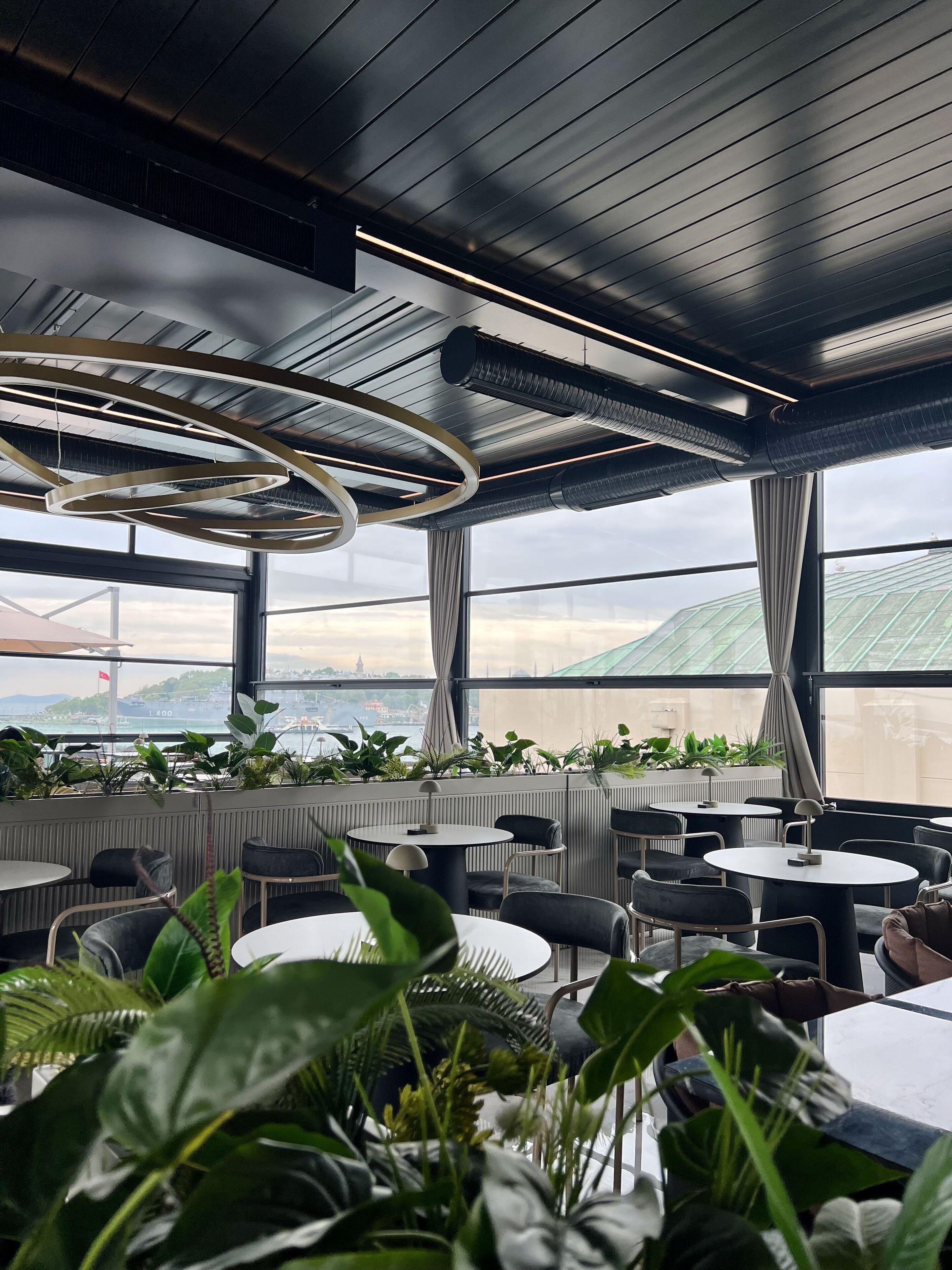 Interior moderno de restaurante con mesas redondas, sillas y ventanas amplias que ofrecen vistas a la ciudad y áreas verdes.