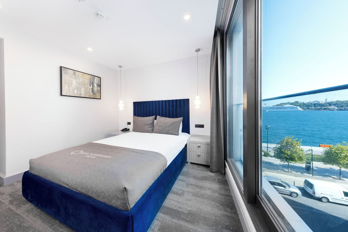 A modern hotel room with a large bed, a view of the sea, and a cityscape in the background.