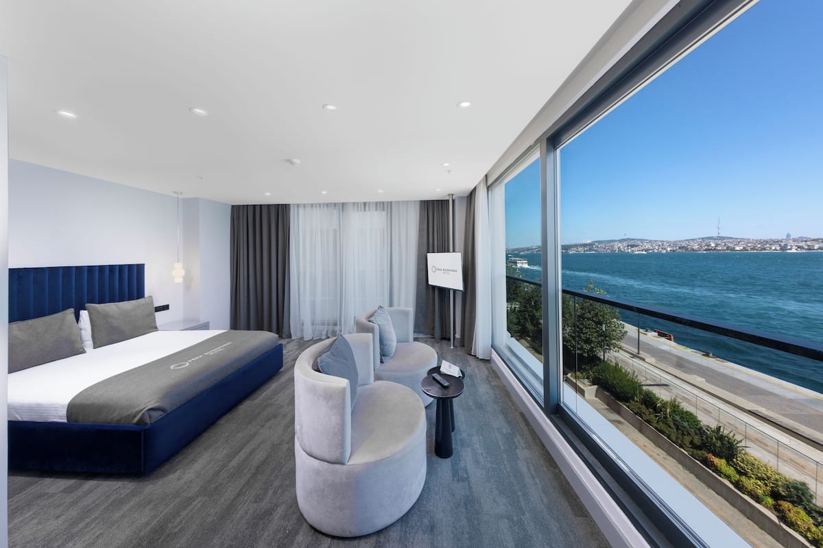 A modern hotel room with a bed, seating area, and a large window offering a view of the sea and cityscape.