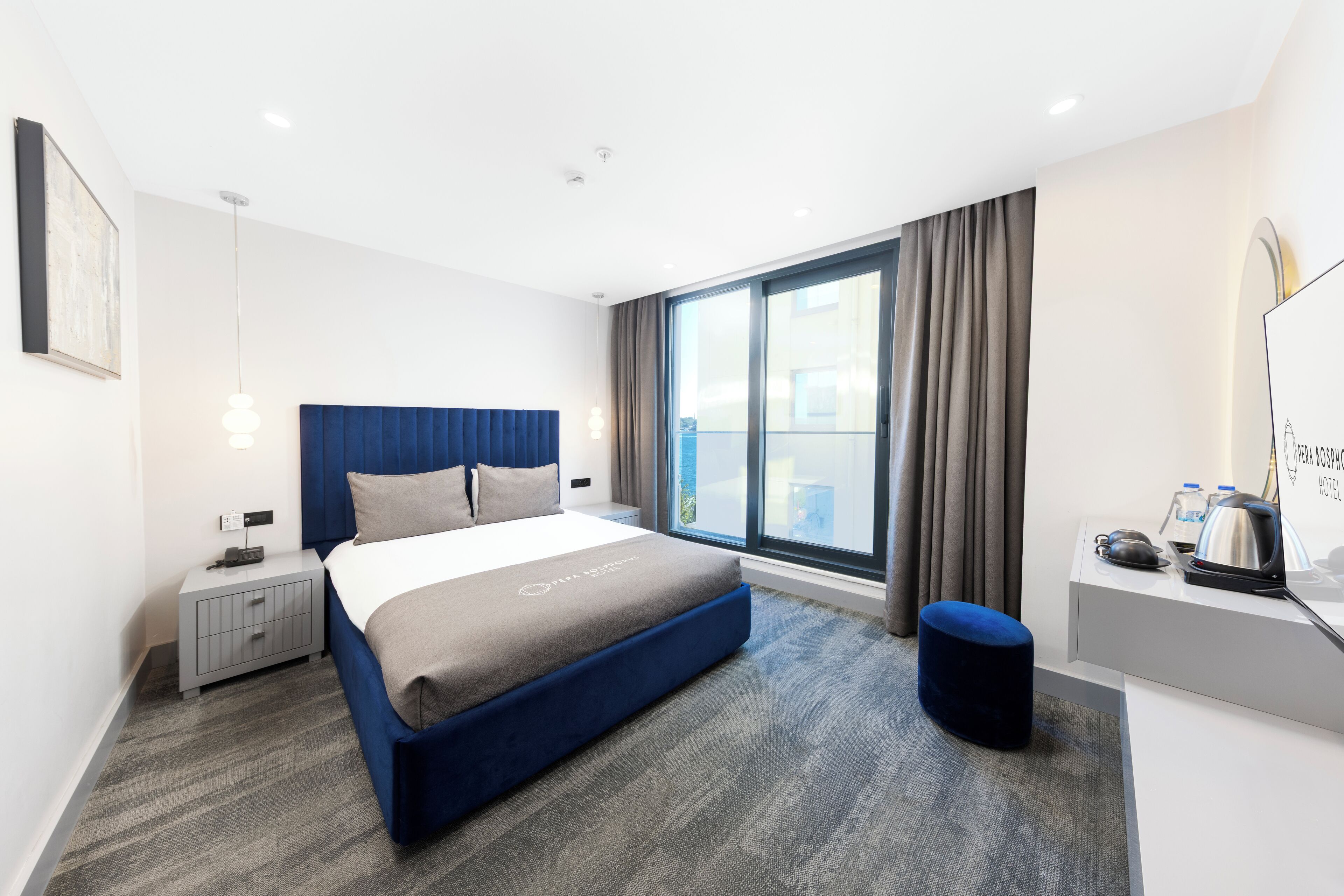 A modern hotel room with a large bed, a desk with a kettle, and a view of the city through the window.