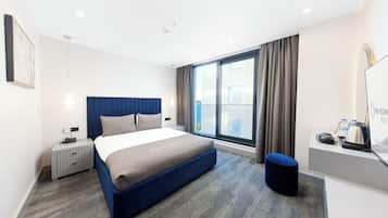 A modern hotel room with a large bed, a desk with a kettle, and a view of the city through the window.